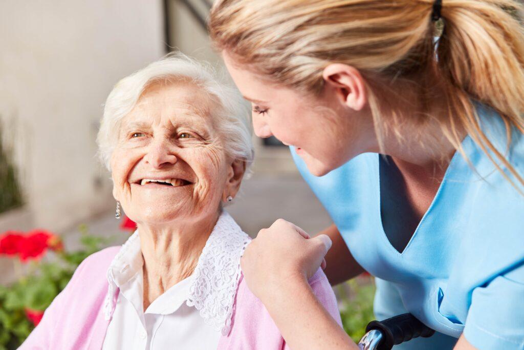 Eine freundliche Pflegekraft in blauer Uniform beugt sich zu einer älteren Frau in einem rosa Pullover und lächelt sie an. Die ältere Frau lächelt ebenfalls und wirkt glücklich.