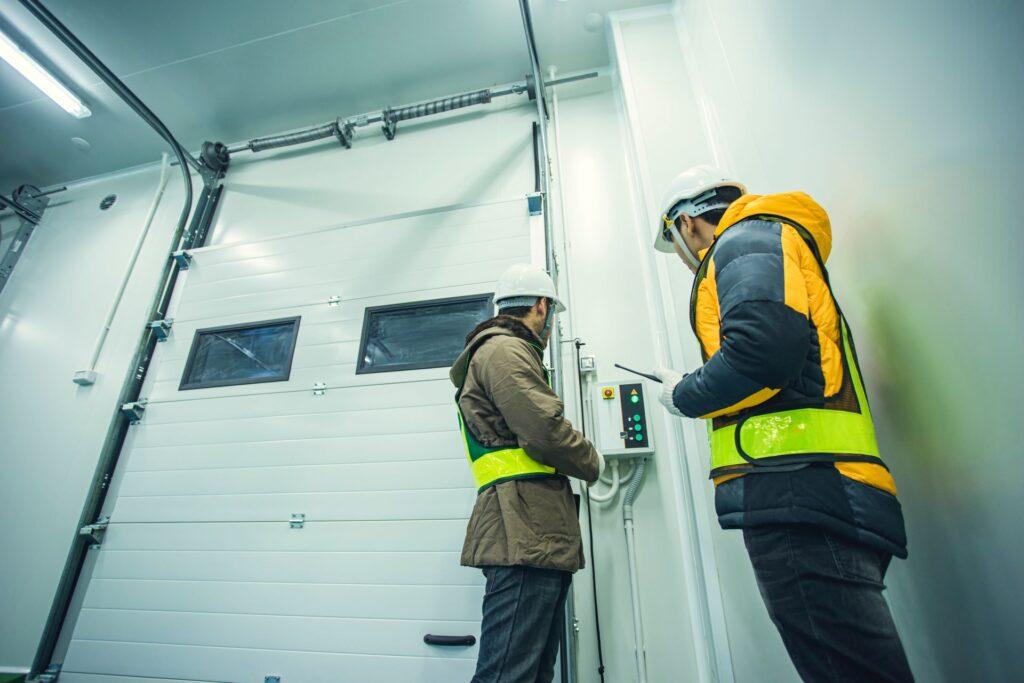 Zwei Männer mit weißen Schutzhelmen und reflektierenden Sicherheitswesten stehen vor einem großen weißen Industrietor, arbeiten an einem Steuergerät, mit einer Zimmertür mit Zarge in einem gut beleuchteten, modernen Industriestandort.